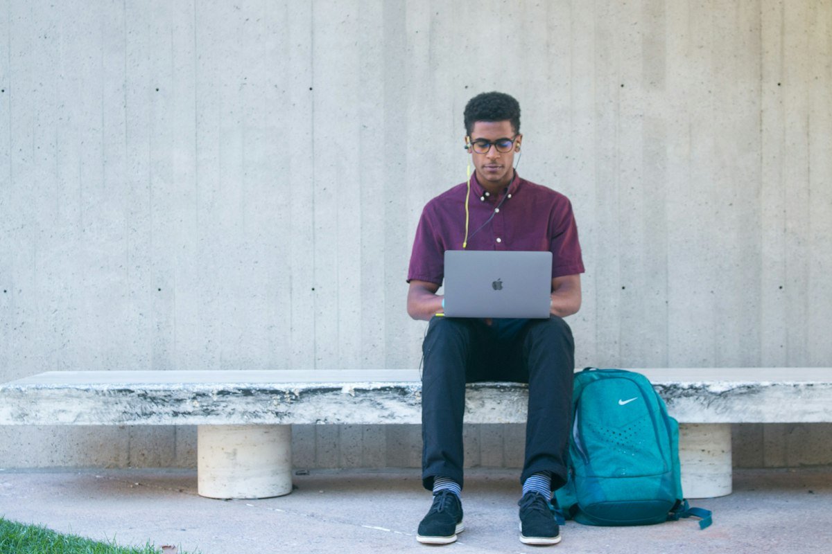 Student using a laptop on a bench with a backpack beside him
